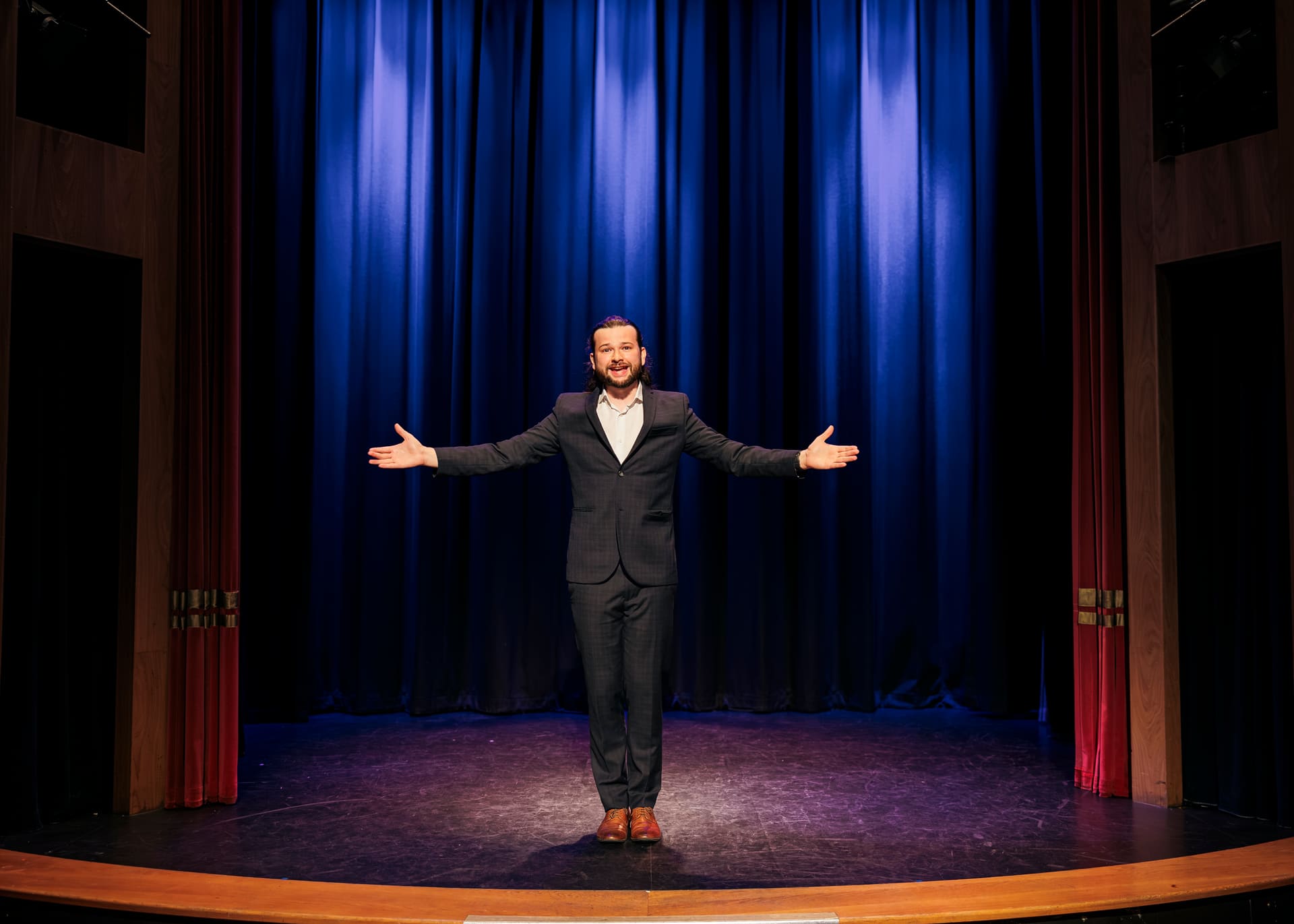 Magician Callum McClure on stage with his arms outstretched in front of a blue theatre curtain