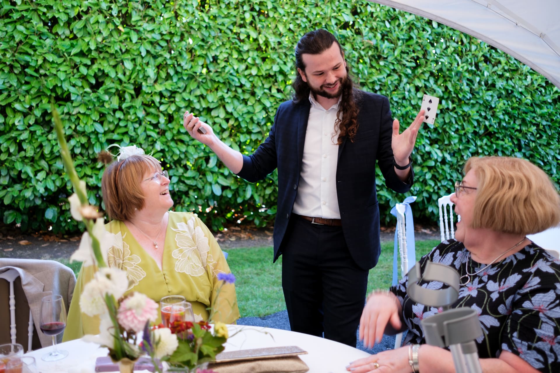 Magician Callum McClure revealing a playing card to two seated guests at an outdoor wedding reception